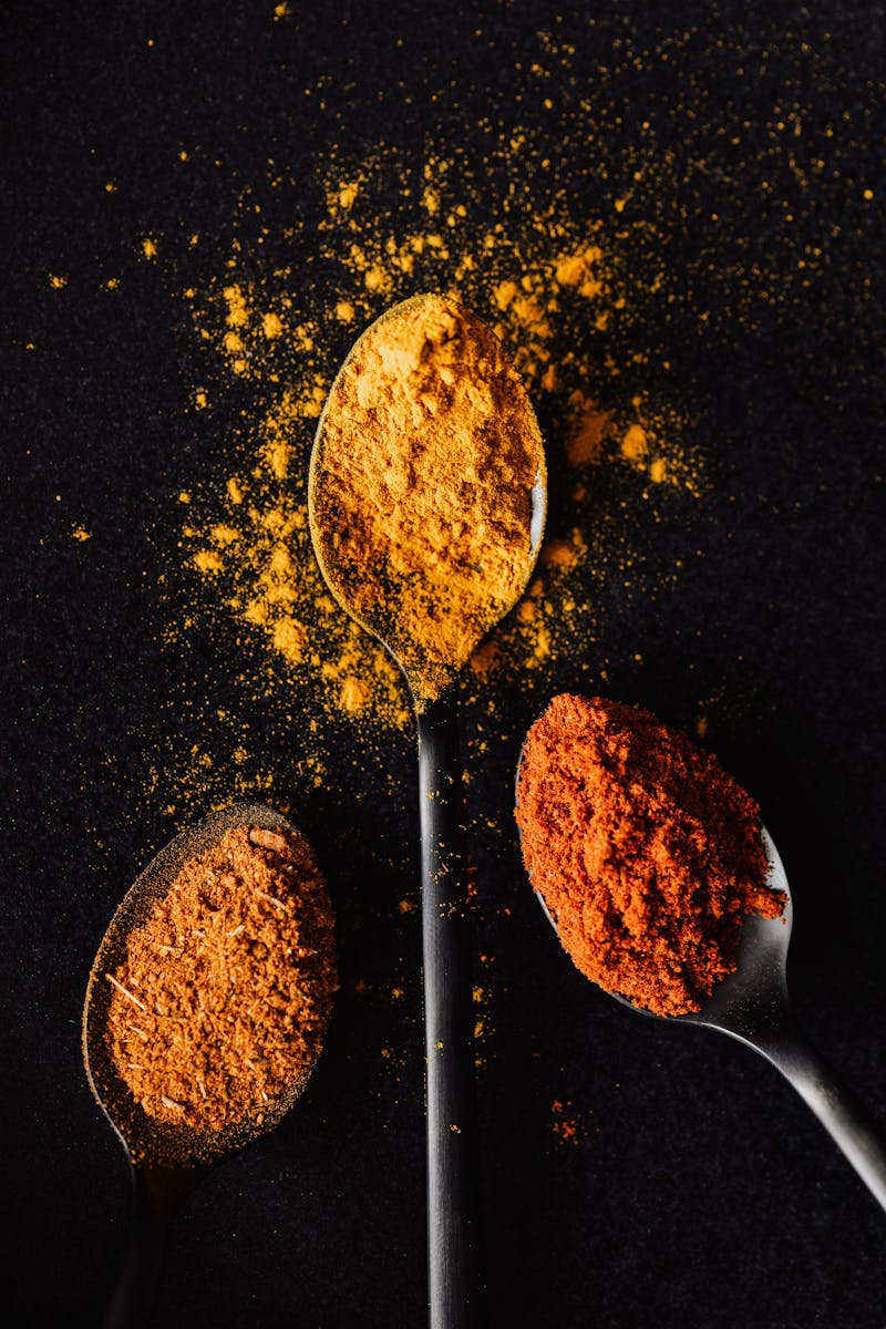Vivid spices in spoons on black background, showcasing turmeric, paprika, and chili powder.